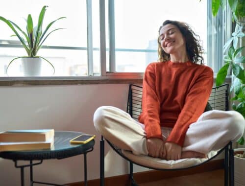 La femme est une jeune femme souriante et décontractée, assise sur une chaise à la maison. La femme qui se sent heureuse et positive se réjouit de son bien-être et se détend près d'une fenêtre dans un appartement moderne et confortable.