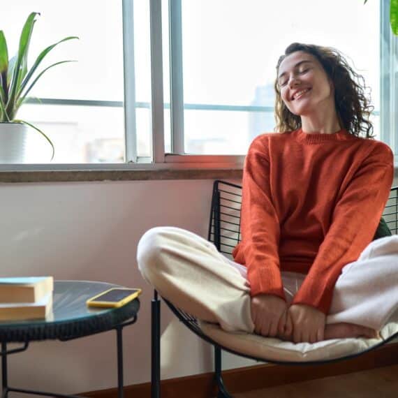 La femme est une jeune femme souriante et décontractée, assise sur une chaise à la maison. La femme qui se sent heureuse et positive se réjouit de son bien-être et se détend près d'une fenêtre dans un appartement moderne et confortable.